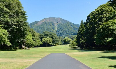Fototapeta premium Paved path through green golf course towards forested mountain under blue sky.