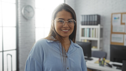 Young woman smiling in an office with a bookshelf and computers in the background, showcasing a bright well-lit modern workplace