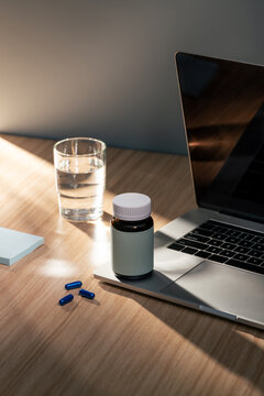 A sunlit wooden desk with a glass of water, blue capsules