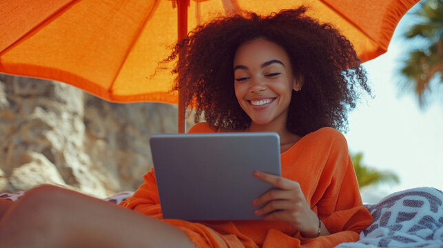 Beautiful smiling young woman lying on sunbed on the beach reading a book on tablet under the parasol