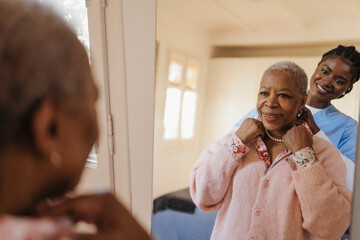 Caregiver helping elderly woman putting necklace in front of mirror