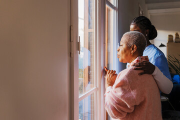 Caregiver comforting elderly woman looking out window