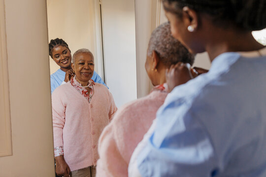 Caregiver helping elderly woman putting necklace in front of mirror - Powered by Adobe