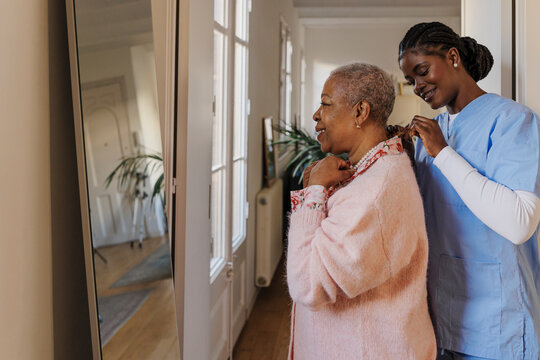 Caregiver helping elderly woman putting necklace in front of mirror