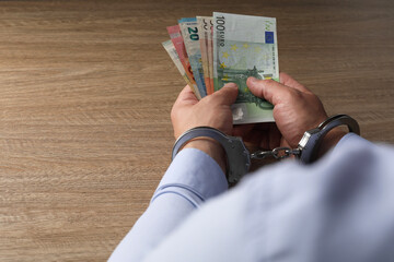 Corruption. Man in handcuffs holding euro banknotes at wooden table, closeup