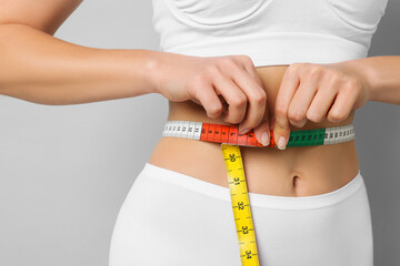Weight loss. Woman measuring her slim waist with tape on light grey background, closeup