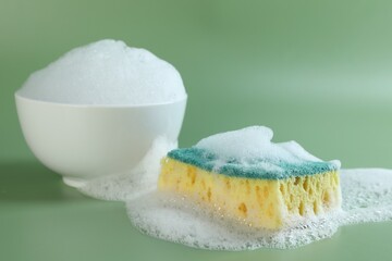 Sponge with foam, bowl and bubbles on green background, closeup