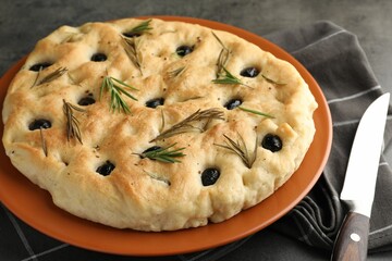 Delicious focaccia bread with olives, rosemary and knife on grey table, closeup