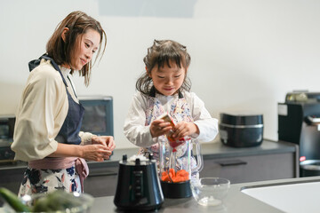 A Japanese parent and child, a woman in her 30s, a man in his 20s, and a 6-year-old girl, are making juice by putting cut carrots in a blender in their home kitchen.