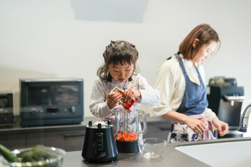 A Japanese parent and child, a woman in her 30s, a man in his 20s, and a 6-year-old girl, are making juice by putting cut carrots in a blender in their home kitchen.