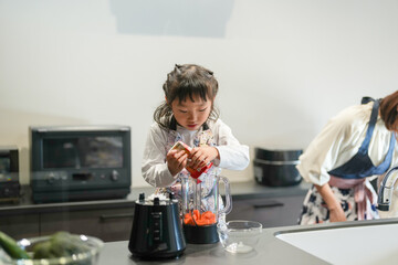 A Japanese parent and child, a woman in her 30s, a man in his 20s, and a 6-year-old girl, are making juice by putting cut carrots in a blender in their home kitchen.