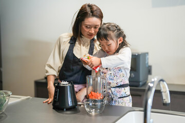 A Japanese parent and child, a woman in her 30s, a man in his 20s, and a 6-year-old girl, are making juice by putting cut carrots in a blender in their home kitchen.