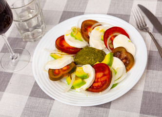 Appetizing caprese avocado salad of sliced ripe tomatoes with creamy mozzarella, onion dressed with fragrant sauce pesto..