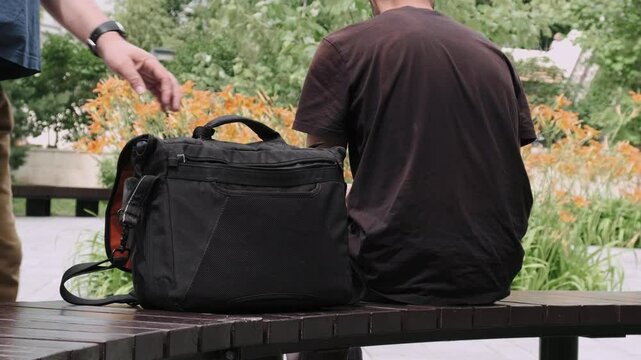 Man with black bag sitting on bench outside while thief discreetly takes out smartphone from it and leaves. Pickpocket robber is stealing phone from handbag. Outdoor crime or stolen property.