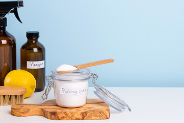 A jar filled with baking soda and a wooden spoon rests on a countertop alongside a bottle of vinegar, fresh lemon, brush and cleaning sponges