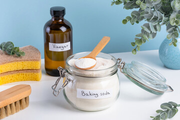 A jar filled with baking soda and a wooden spoon rests on a countertop alongside a bottle of vinegar and cleaning sponges. DIY cleaning method using natural products to remove stains. top view 