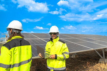 Two technicians in safety gear and reflective jackets inspect a solar panel installation at a solar farm. They are engaged in maintenance and operational checks under a clear blue sky.