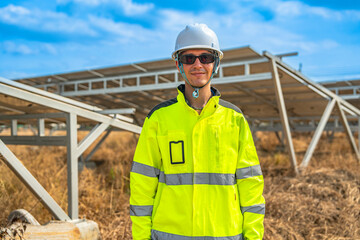 Smiling solar energy engineer in a reflective jacket and helmet stands confidently with arms crossed at a solar farm. Photovoltaic panels are visible in the background under a clear blue sky.