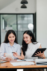 Two women working together in a modern office, using digital devices and discussing documents.