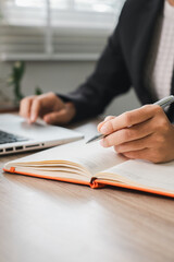 Close-up of a business professional writing in a notebook while using a laptop, showcasing productivity and focus.