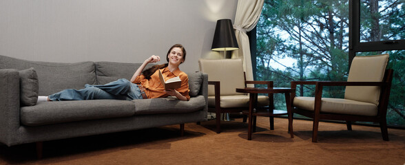 Smiling young woman relaxing on a gray sofa in a cozy living room, enjoying a book with a warm ambiance, perfect for home lifestyle concepts