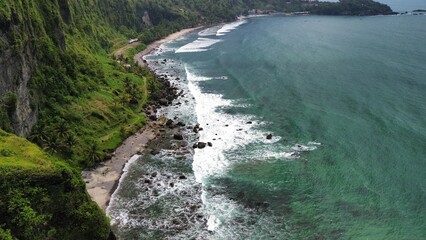 Aerial drone view of coastline with hills and trees, as well as view of coral cliffs and sea with waves from the ocean in Menganti Beach Kebumen Central Java Indonesia