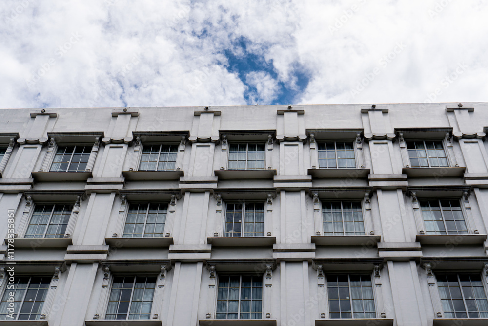 Aufkleber a symmetrical view of a gray building with repeating windows, against a cloudy sky backdrop.