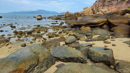 A rocky coastal landscape with scattered boulders in Hon Chong cape rocks garden on the Nha Trang beach, Vietnam with a calm sea and mountains in background.