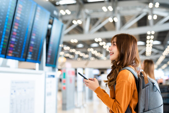 Young asian woman in international airport, using mobile smartphone and checking flight at the flight information board