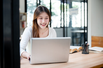 Fototapeta premium A smiling, happy Asian female college student working on her project on her laptop in a coffee shop.