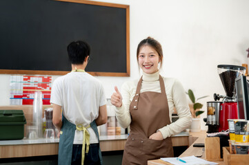 A smiling Asian female barista is giving a thumbs-up to the camera while standing at the counter.