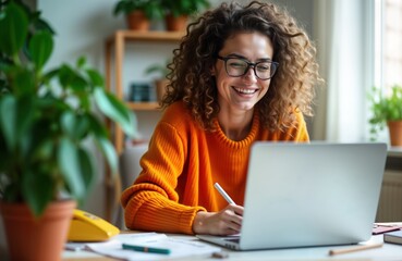 Young woman sits at home office desk using laptop, taking notes. Happy expression, focused on laptop screen. Likely watching webinar video call. Casual attire. Looks like working remotely studying