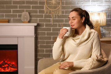 Beautiful young happy woman with cup of hot chocolate sitting on armchair near fireplace in evening at home