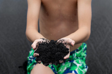 A teen boy in vivid swimming trunks is sitting in the beach and holding a handful of unique beautiful black volcanic sand made of lava. Black sand beach, Big Island, Hawaii, USA.