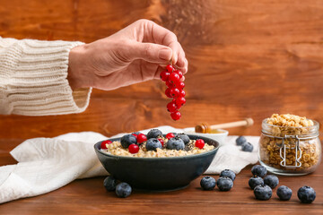 Young woman adding cranberry in bowl of tasty oatmeal on wooden background