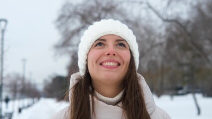 Portrait of a woman in a warm white knitted hat outdoors in winter. Young woman smiling and enjoying the beautiful weather and the first snow, laughing and looking at the sky. Active winter recreation