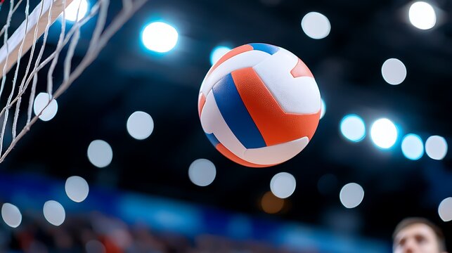Close-up of a volleyball approaching a net during an indoor match, with blurred background lights and spectators.