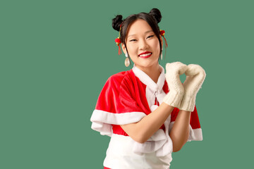 Beautiful Asian woman in Santa costume showing heart shape with her hands on green background