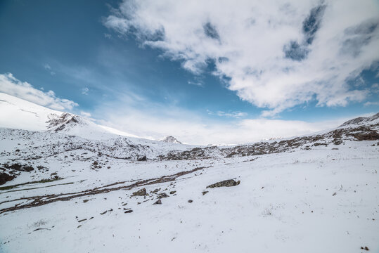 Cold snow-white landscape with snow-covered pointy peak under clouds in blue sky. Snowy rocky mountainside and mountain peaked top. Last year's grass and stones in snow and sharp pointed pinnacle.