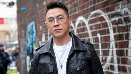Fototapeta premium Urban male with leather jacket, stylish glasses, and cross necklace posing against graffiti brick wall, modern streetwear, confident expression.