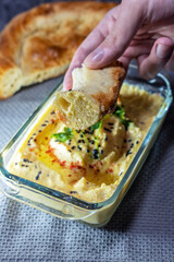 A man's hand holds a flatbread dipped in hummus, which is sprinkled with parsley, paprika and black sesame seeds and drizzled with olive oil, vertical photo.