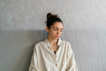A woman with a bun is sitting on a grey sofa, holding her face in her hand, expressing sadness and...