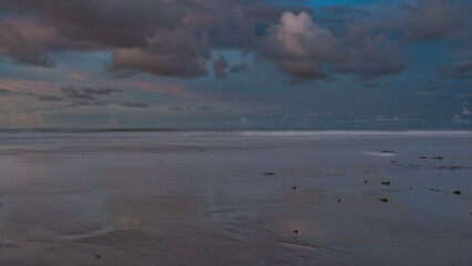Evening by the ocean. Pastel colors. The clouds in the blue sky are highlighted in pink. The waves are foaming, spreading on the beach. Bunches of algae on wet sand. Long exposure. Malaysia. Borneo. 