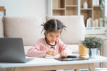 An Asian female and her daughter sit on the sofa in the living room during the day, using a laptop, headphones, and mobile device while studying at the desk with books around them.