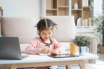 An Asian female and her daughter sit on the sofa in the living room during the day, using a laptop, headphones, and mobile device while studying at the desk with books around them.