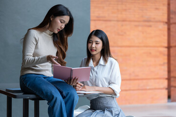 Two young asian businesswomen collaborating on a project, explaining concepts from a pink notebook while taking notes, highlighting teamwork in a modern office