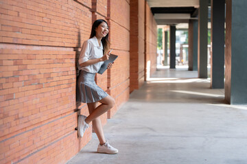 Young asian student is smiling while leaning against a brick wall in a university hallway, holding a book and a pen, enjoying her break time