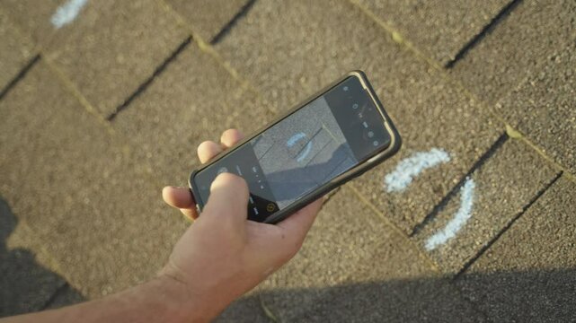 Roofer photographing marked hail damage on roof shingles using a smartphone for insurance evaluation.