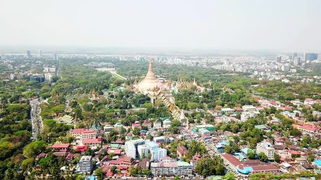 Drone capturing Shwedagon Pagoda, a magnificent golden stupa, surrounded by lush greenery and urban development in Yangon Rangun, Myanmar, showcasing the city's blend of religious and modern life.