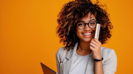 A young woman with curly hair smiling while talking on her phone and holding a laptop, posed against a vibrant orange background.

